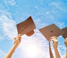 Four hands holding graduation hats on background of blue sky.